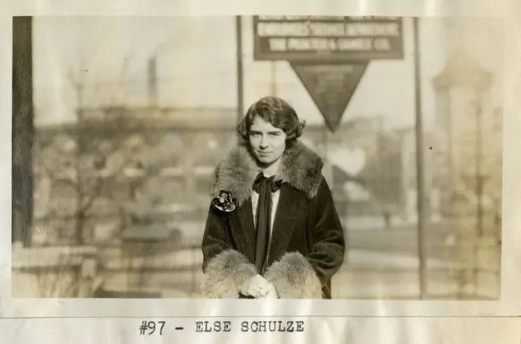 A white woman with short hair wears a coat with fur trim on its collar and wrist areas. In the black and white photo, she stands outside in front of a building.