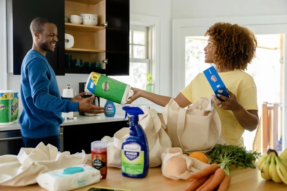 Two people are in a kitchen, organizing groceries and cleaning supplies. One person passes a roll of Bounty paper towels to the other.