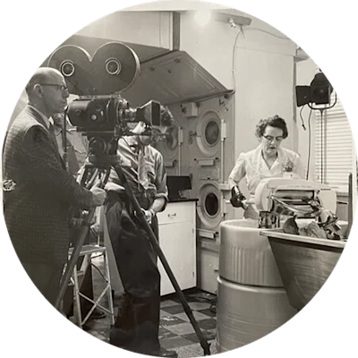 A woman in a white uniform operates a mid-century mechanical laundry machine. Two men stand behind a camera tripod as they film her doing laundry. The image is in black and white.