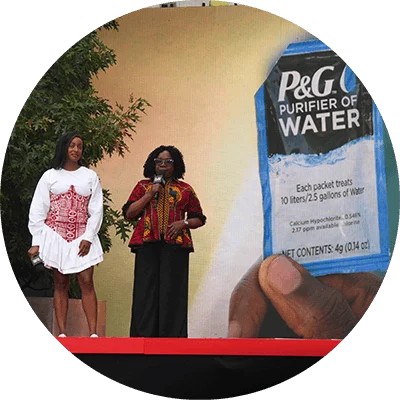 Two black women on stage. One is in a white dress and the other in colorful attire. They speak in front of a large image of a P&G Purifier of Water packet.