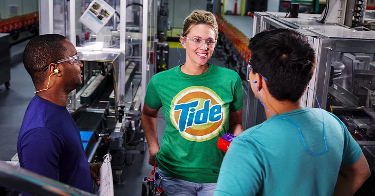 Three P&G plant employees next to a Tide manufacturing line. The female employee facing the camera is wearing a Tide t-shirt.