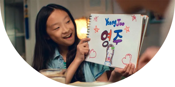 A girl smiles and holds up a drawing of herself and her name "Yeong Joo"