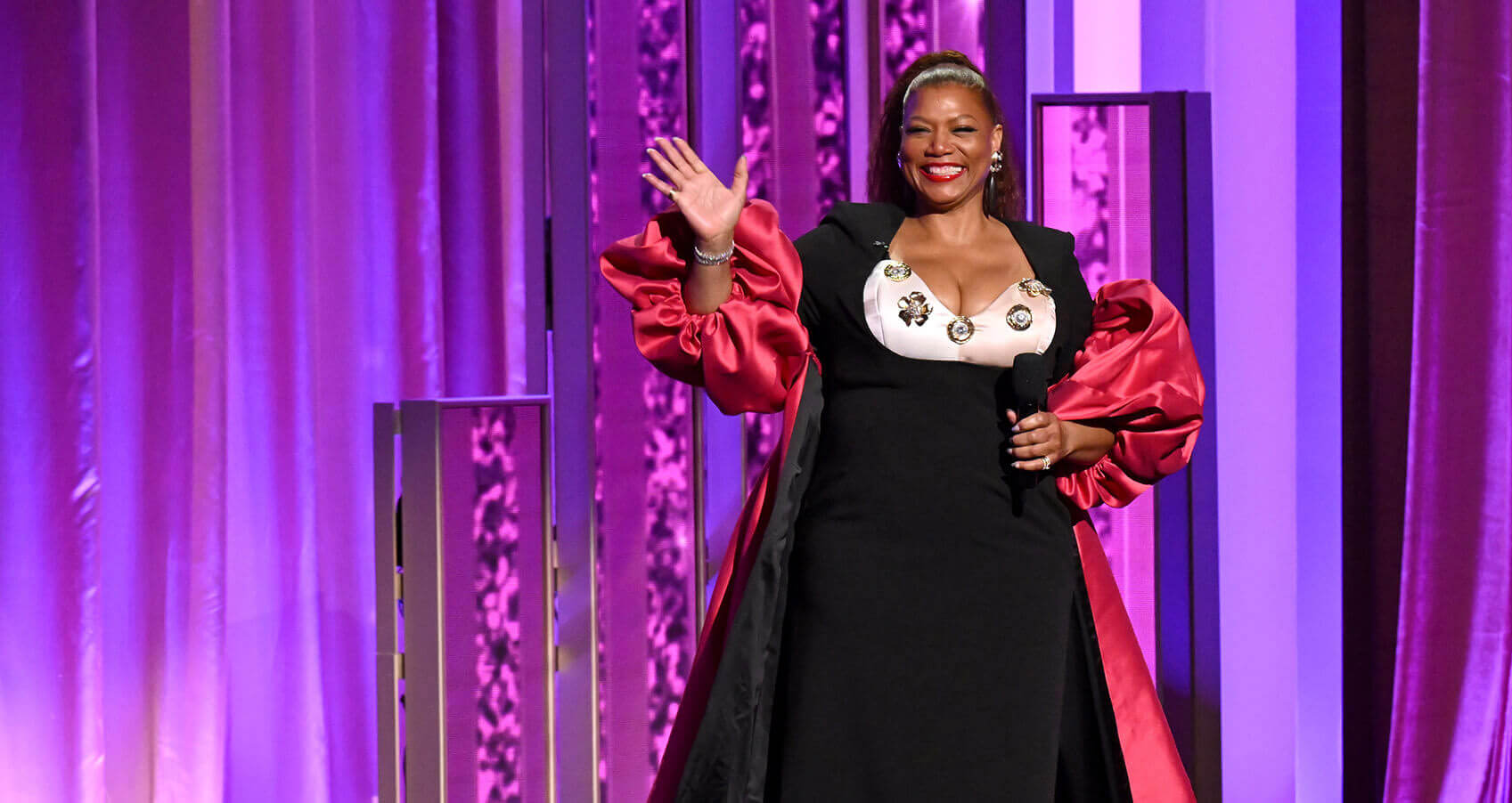 A Black woman in a formal black dress stands on stage with a microphone in her hand, smiling and waving at the camera.