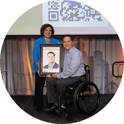 A black woman with short dark curly hair. A white man sits in a wheelchair next to her.  They hold up a portrait with black text that says “equality & inclusion global inspirational leader.”