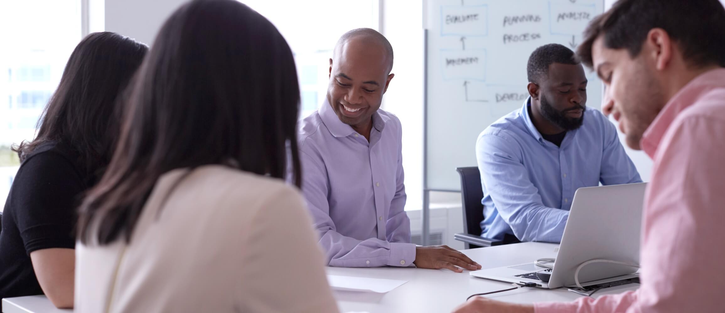 Group of employees works together around a table. Man in center of image smiles while working at laptop  