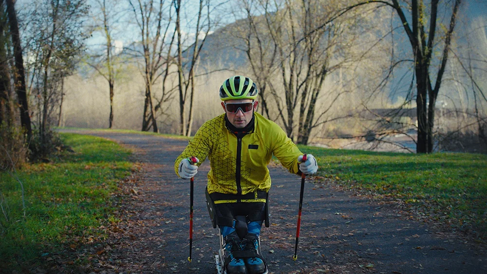 A man in a bright yellow jacket, green helmet, and sunglasses practices Para cross-country skiing on a paved path, holding ski poles and using adaptive equipment. Bare trees line the path, and mountains are visible in the distant background.