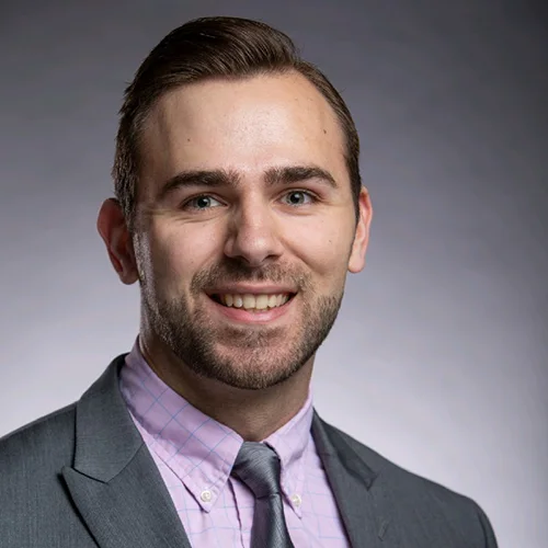 Headshot of a man with light eyes and a friendly smile, dressed in business casual attire against a gray background.
