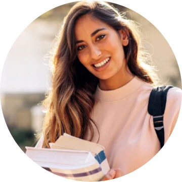 A woman with long brown hair is smiling towards the camera and holding two textbooks. There is a backpack strap on her shoulder.