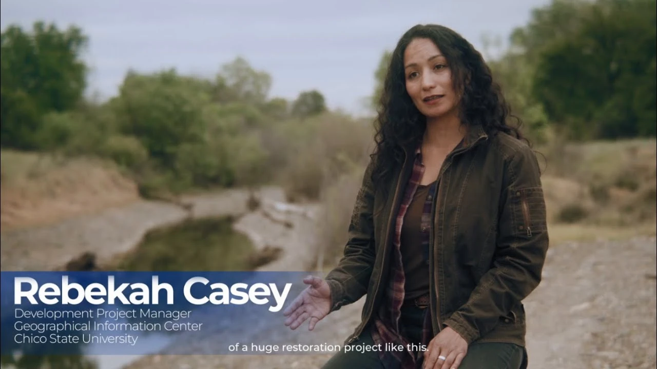 A woman with long dark curly hair. She stands outside in front a dry landscape with green brush.