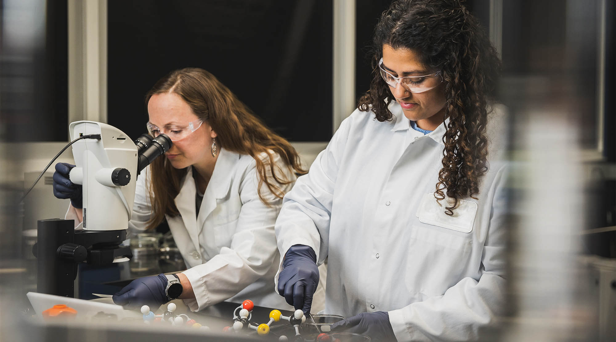 Two female scientists wear white lab coats, gloves, and safety goggles. One of the women is staring into a microscope.