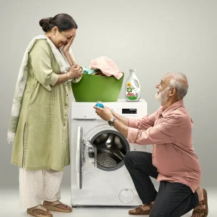 A man and woman are in front of a laundry machine with a basket of laundry and bottle of Ariel laundry detergent on top of it. The man is kneeling in front of the woman proposing. The woman has her hand on her face.