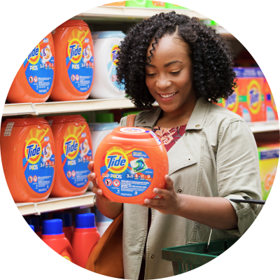 Woman with dark curly hair standing in front of Tide shelf, holding Tide product in her hands and smiling.
