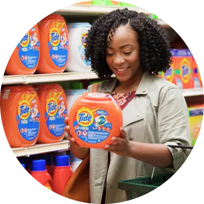Woman with dark curly hair standing in front of Tide shelf, holding Tide product in her hands and smiling.