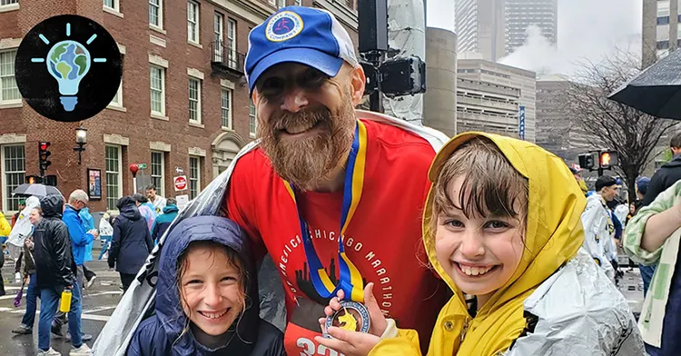 An adult white male and two young girls pose in raincoats as they smile directly at the camera. Additional pedestrians and a city landscape are in the background.