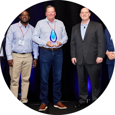 Five men stand side by side on a stage with a dark backdrop. The man in the center holds up a glass trophy.