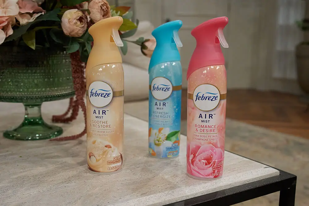 Three colorful spray cans of Febreze air freshener. They are placed on an end table, with a vase of flowers in the background.