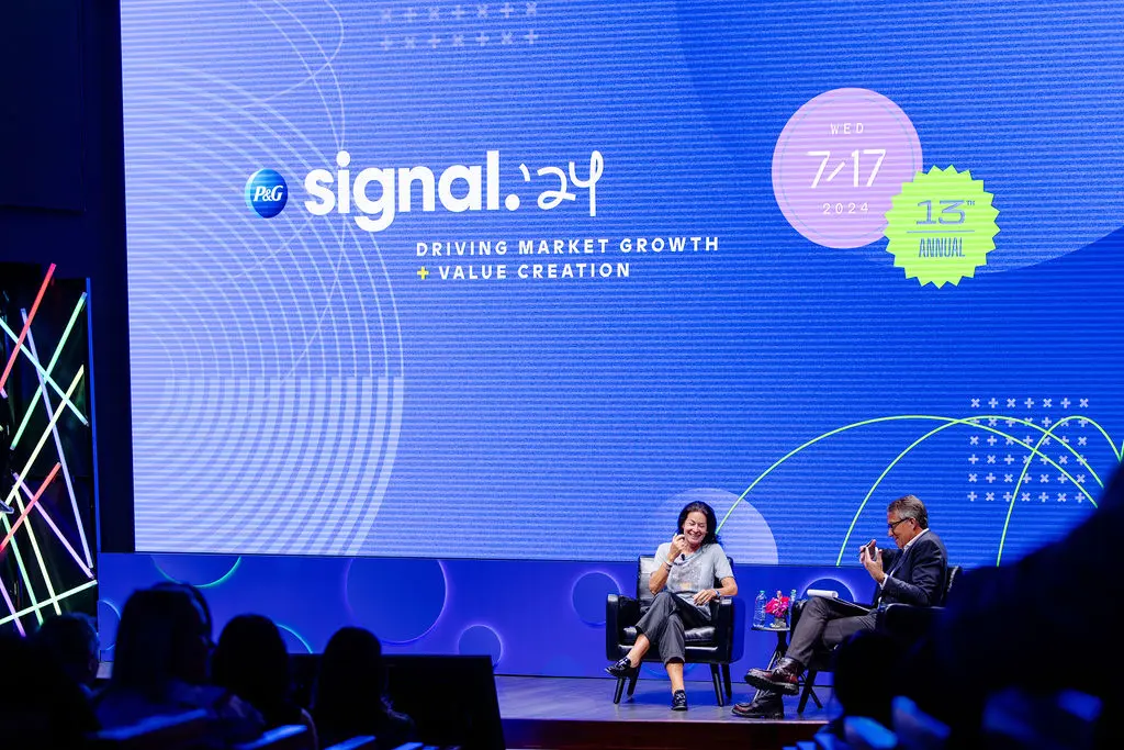 A woman with dark hair and a white man sit and talk on a stage. A large blue graphic is displayed in the background with text that says "Signal '24. Driving market growth and value creation."