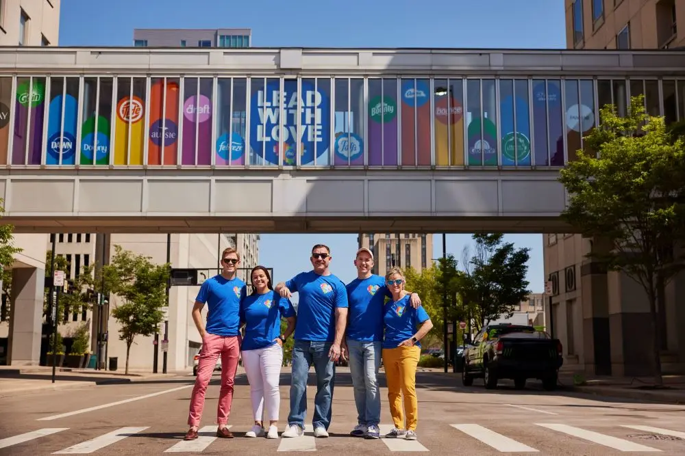 Five P&G employees in matching blue shirts smile and stand on the street under a bridge decorated with rainbow colors.