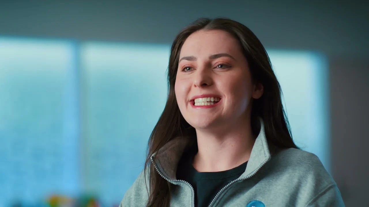 Smiling woman with long dark hair wearing a light gray zip-up jacket in a softly lit office setting.