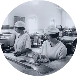Two young women in white caps and uniforms. They stand in a manufacturing facility behind a long table with a long row of soap bars. The photo is from 1910.