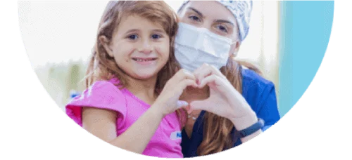 A woman who is a professional dentists is wearing a mask and scrubs sitting next to a young girl with a smile. They put their hands together to create a heart symbol
