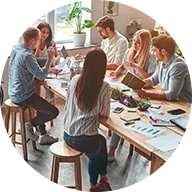 A group of six people engaged in a discussion sit around a wooden table with papers, notebooks, and electronic devices spread out. A blue circle outline surrounds the icon.