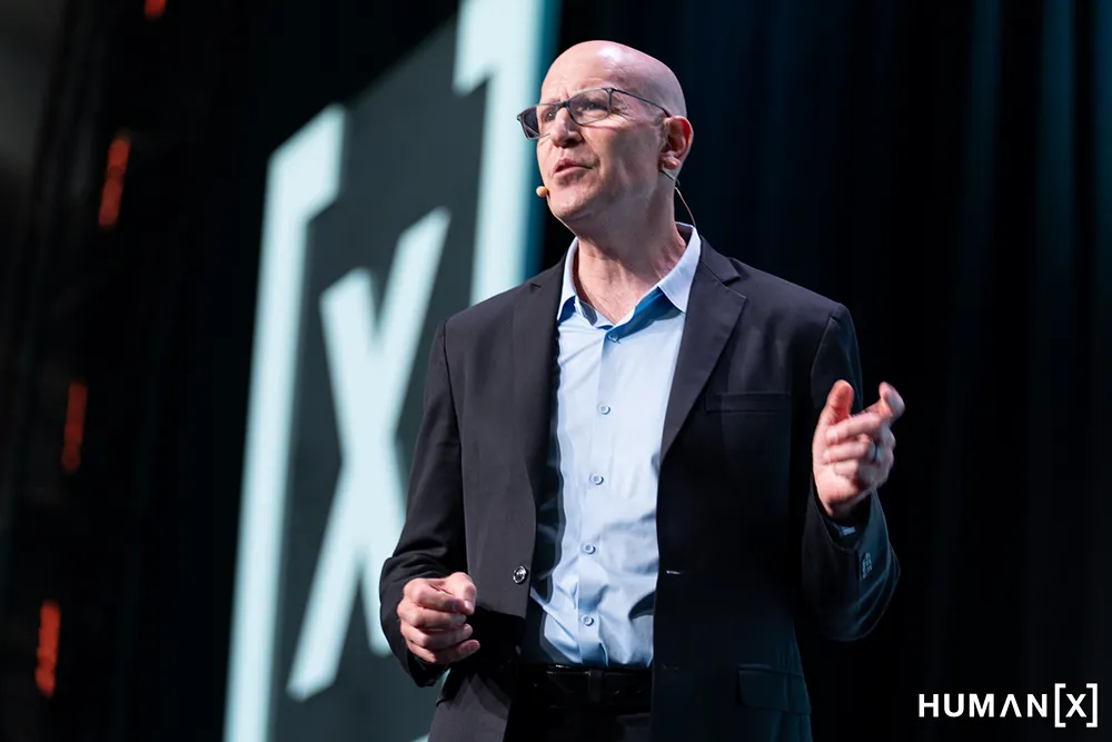 A man in a light blue shirt and black suit jacket. He is wearing a microphone and speaking on a stage.