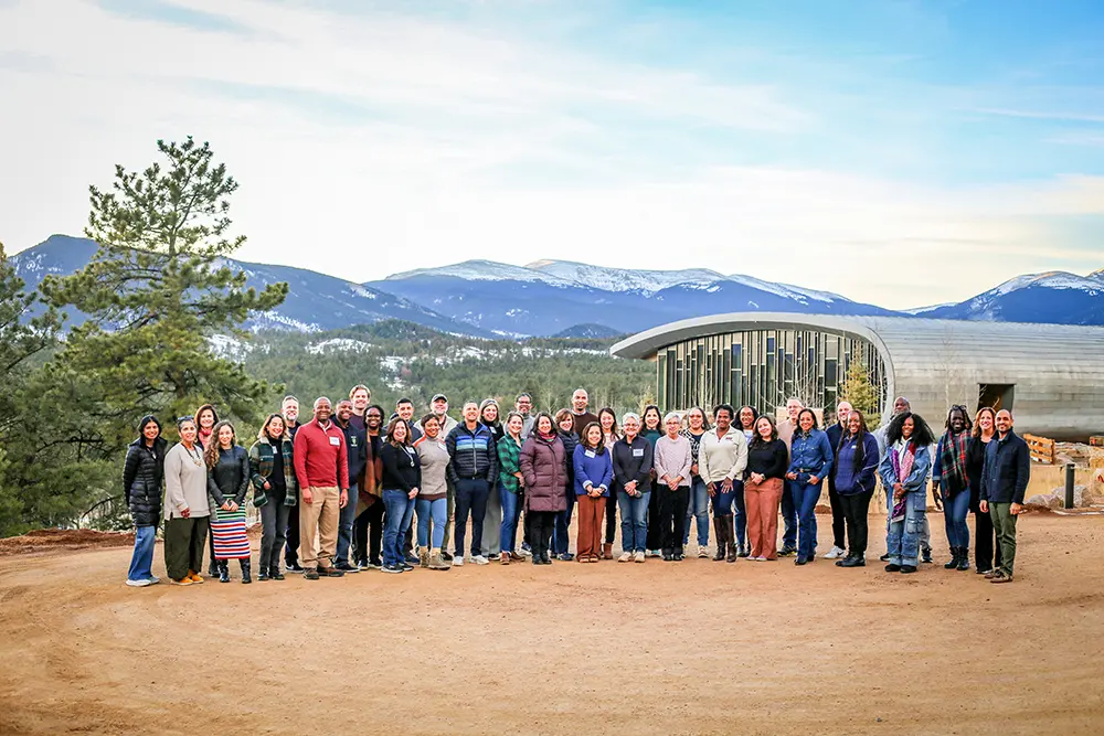 A large group of people stand and pose for a photo amidst the backdrop of snowcapped mountains, trees and an oval shaped, silver building.
