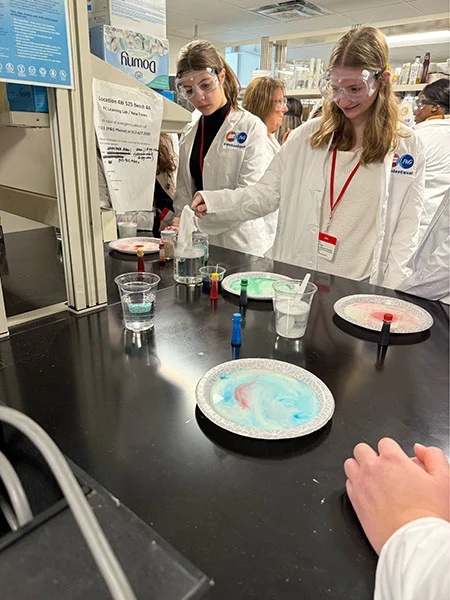 Two young women wear lab goggles and white lab coats. They stand over a black table and hold a white cloth over a beaker filled with liquid. Paper plates holding colored liquids sit on the counter.