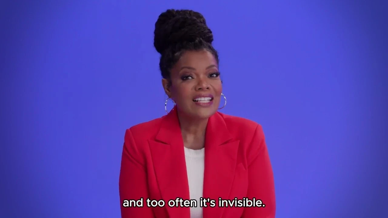 In this video, a Black woman wearing a red blazer and gold hoop earrings and with her hair tied up in a bun talks to the camera about caregiving.