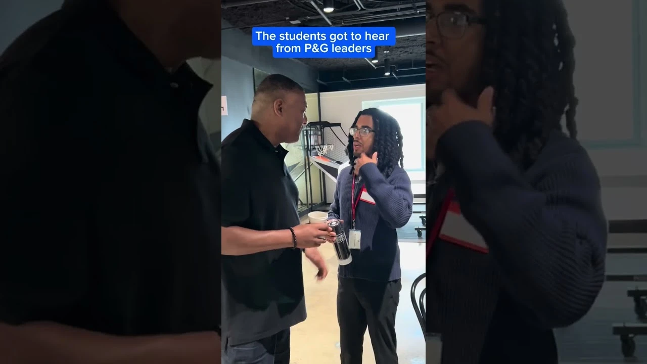 Two people talking at an indoor event space, one holding a drink, with a screen in the background and on-screen text that reads ‘The students got to hear from P&G leaders.