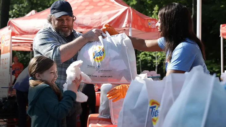 A woman in a light blue t-shirt is handing a man in a black baseball cap and plaid shirt a white tote bag with the Tide logo on it. There is a young girl next to him holding a white stuffed bunny. Tote bags are on the table and an orange tent behind them. 