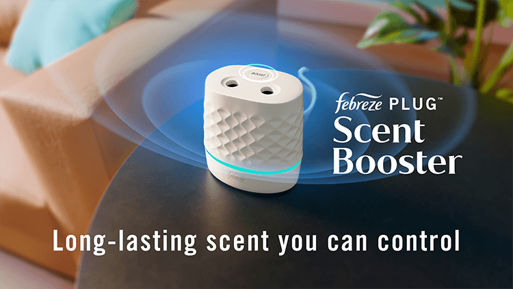 A white oval shaped device sits on a small end table. Two holes and a boost button are at the top of the device. White text says “Febreze plug scent booster. Long lasting scent you can control.”