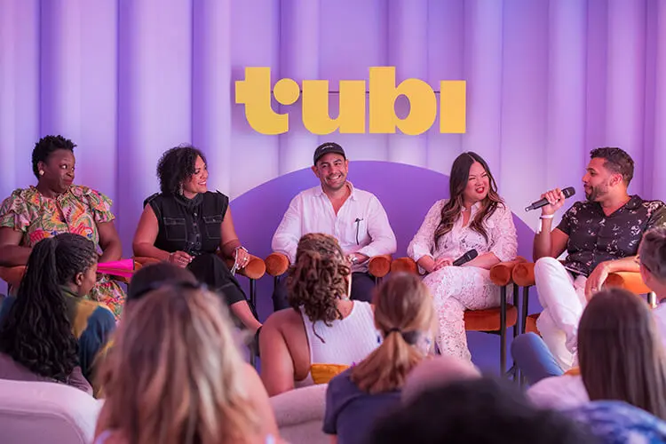 A group of diverse men and women sit together on a stage, engaged in conversation. Yellow letters that say "tubi" hang above them on a purple backdrop.