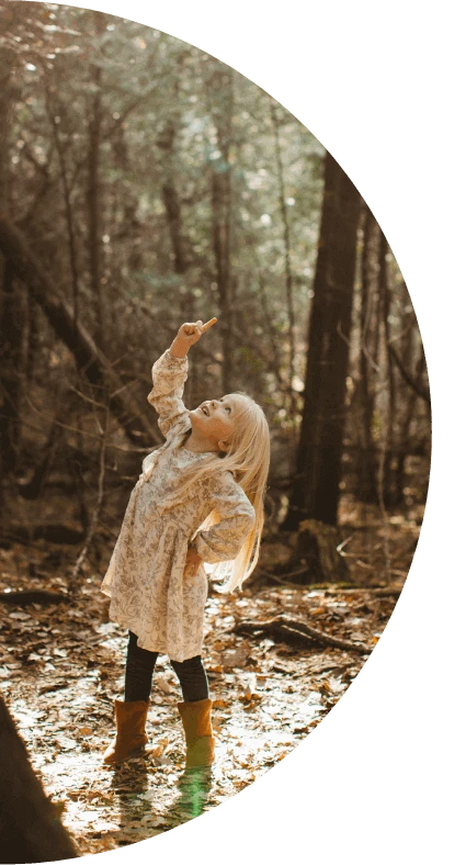 A young girl standing in a wooded area smiles while looking upward and pointing to the sky