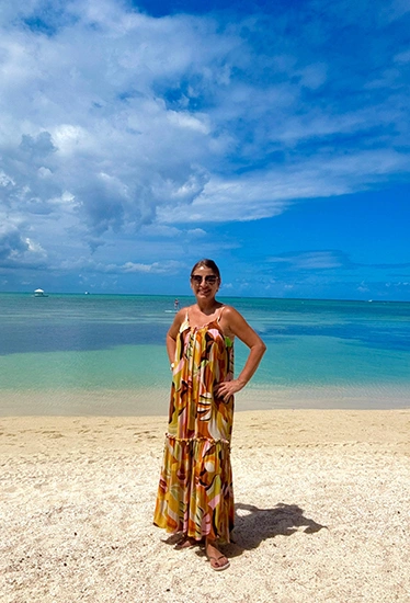 A smiling woman stands on a sunny beach, wearing dark sunglasses and a bright dress.