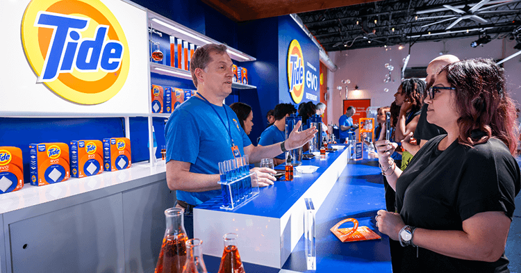 A white man wearing a blue t-shirt stands behind a long booth. The yellow, orange and blue Tide logo and orange colored Tide packaging are displayed behind him as he talks to a woman in a black shirt and glasses.
