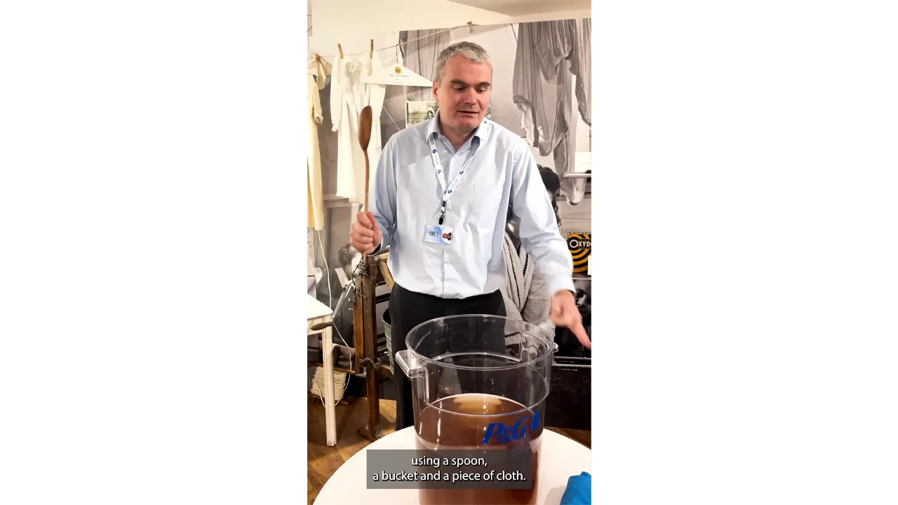 A man in a light blue, long-sleeve button down shirt and lanyard holds a small packet while he stands behind a large clear container filled with murky brown water.