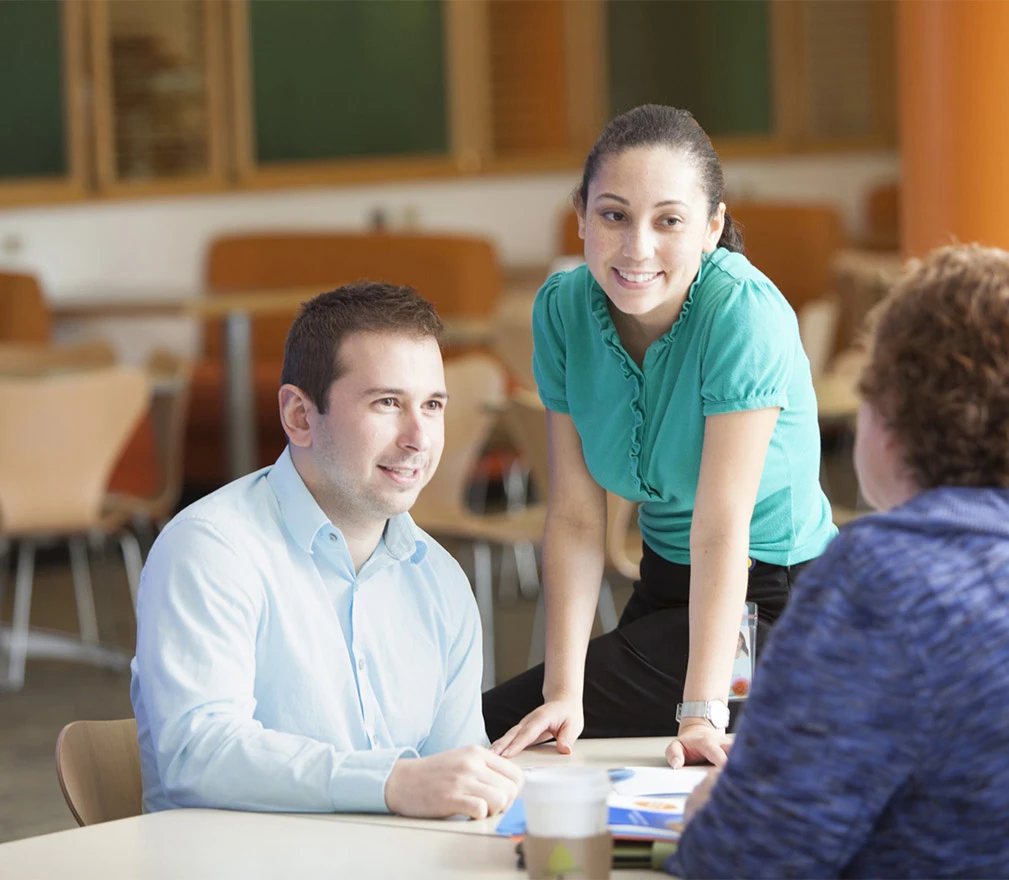 A man in a button-down shirt and a woman in a teal shirt are smiling and listening to another woman speaking to them.