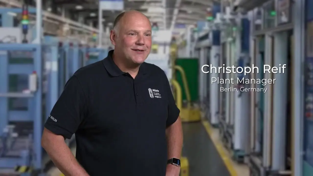 A man in a short sleeve dark shirt. He stands inside a large manufacturing plant.