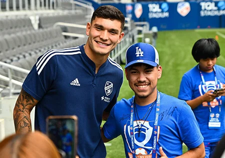 A Hispanic male in a dark blue soccer uniform. He stands next to a young Hispanic male student in a bright blue hat and t-shirt. They are standing on a soccer field.