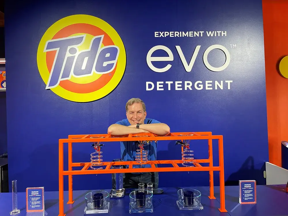 A smiling man leans over an orange science display with glass tubes and beakers in front of a blue wall reading "Experiment with Tide evo Detergent."