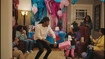 A Black man and little girl dance in the middle of a living room. Several women are sitting in the background. Blue and pink decorations adorn the room.