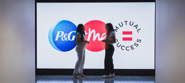 Two women face each other as they engage in conversation. They are standing at the end of a hallway, in front of a digital display of a blue, white and pink logo.