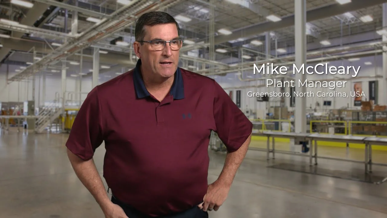 A white man with glasses and a dark red shirt. He stands inside a manufacturing plant.