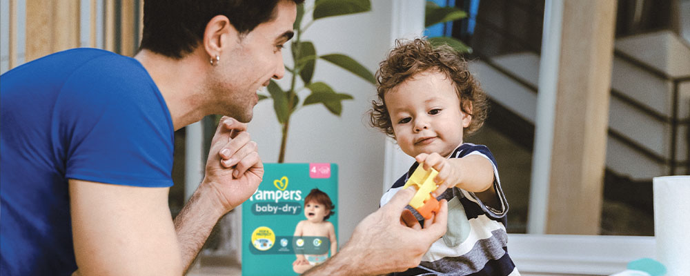 Man on floor playing with baby, handing the baby a toy car. A pack of Pampers diapers is in the background.  