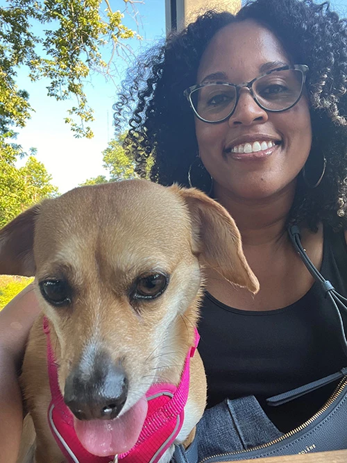 A black woman with dark, curly shoulder length hair and glasses sits next to her dog. She is smiling directly at the camera.