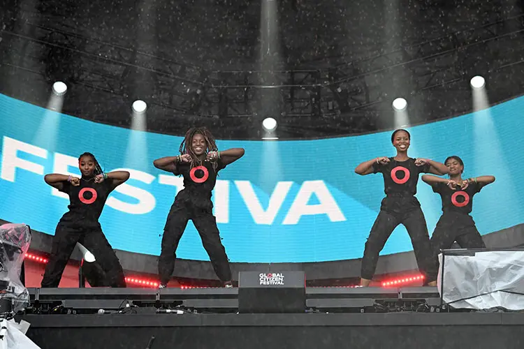 Four young Black women are dancing on a large outdoor stage. They're smiling and wearing matching black activewear outfits that feature a large red circle on the front of their t-shirts.