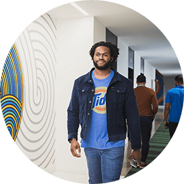 A young man in a t-shirt, jacket, and jeans walks through the hallways of his office.