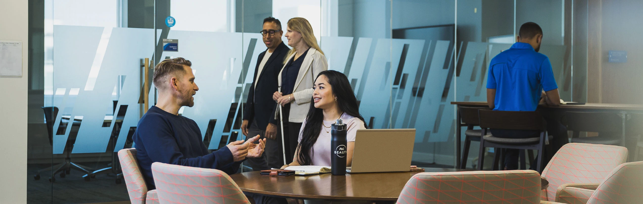 A man and woman are working together at a conference table, talking together. Another man and woman are walking outside the glass-walled room, and another man works at a separate table, his back to the others.